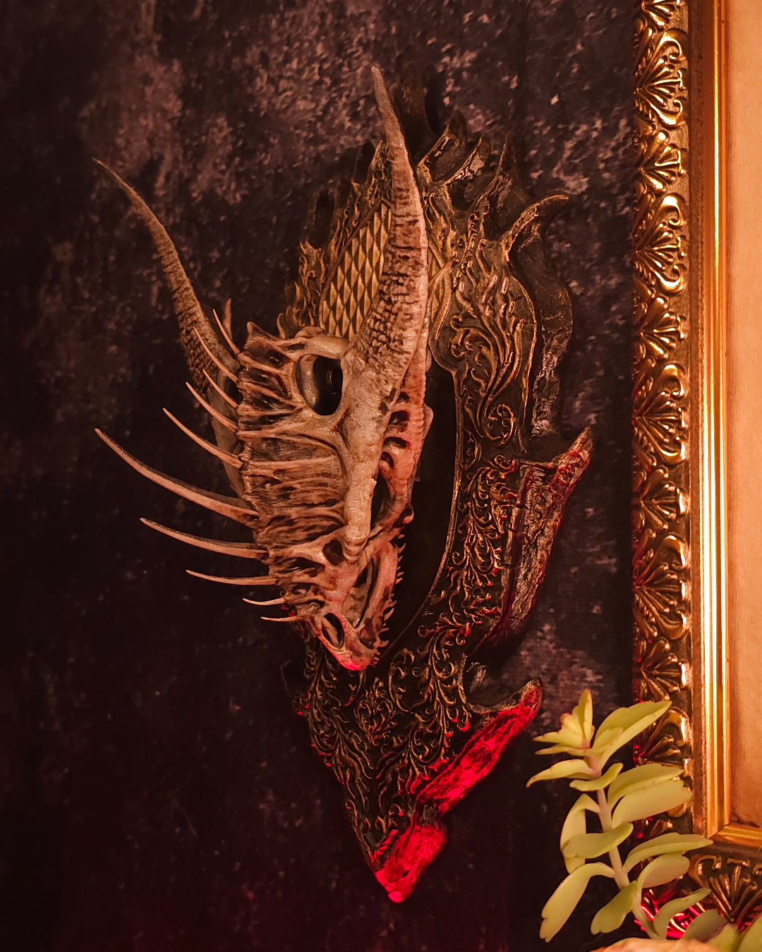 Trophée mural représentant un crâne de dragon avec longues cornes et détails sculptés, fixé sur un support ornemental noir et doré avec éclairage rouge dramatique.