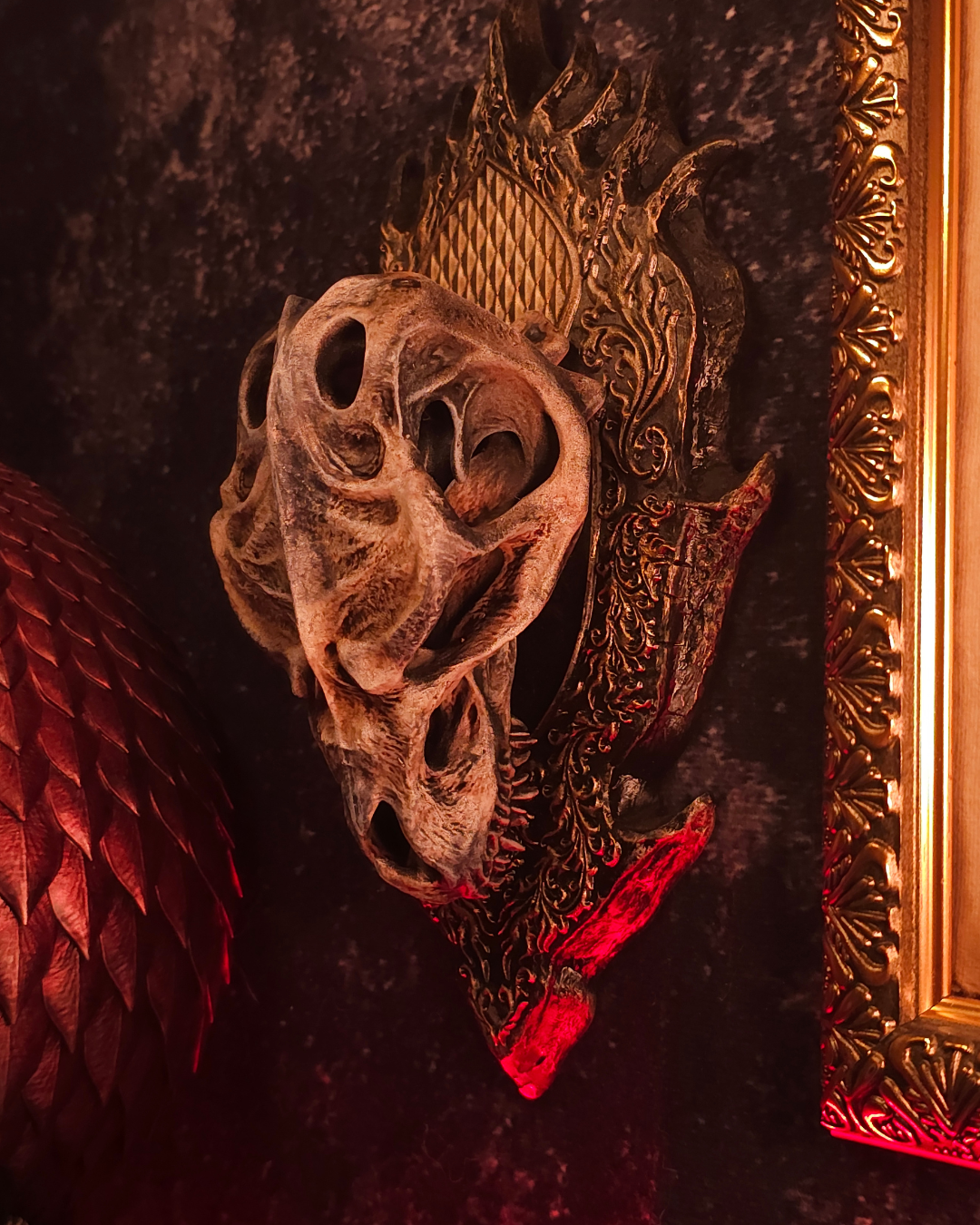 Crâne de dragon godzilla large avec orbites ouvertes et mâchoire détaillée, fixé sur un support sculpté noir avec lumière rouge et décor gothique.