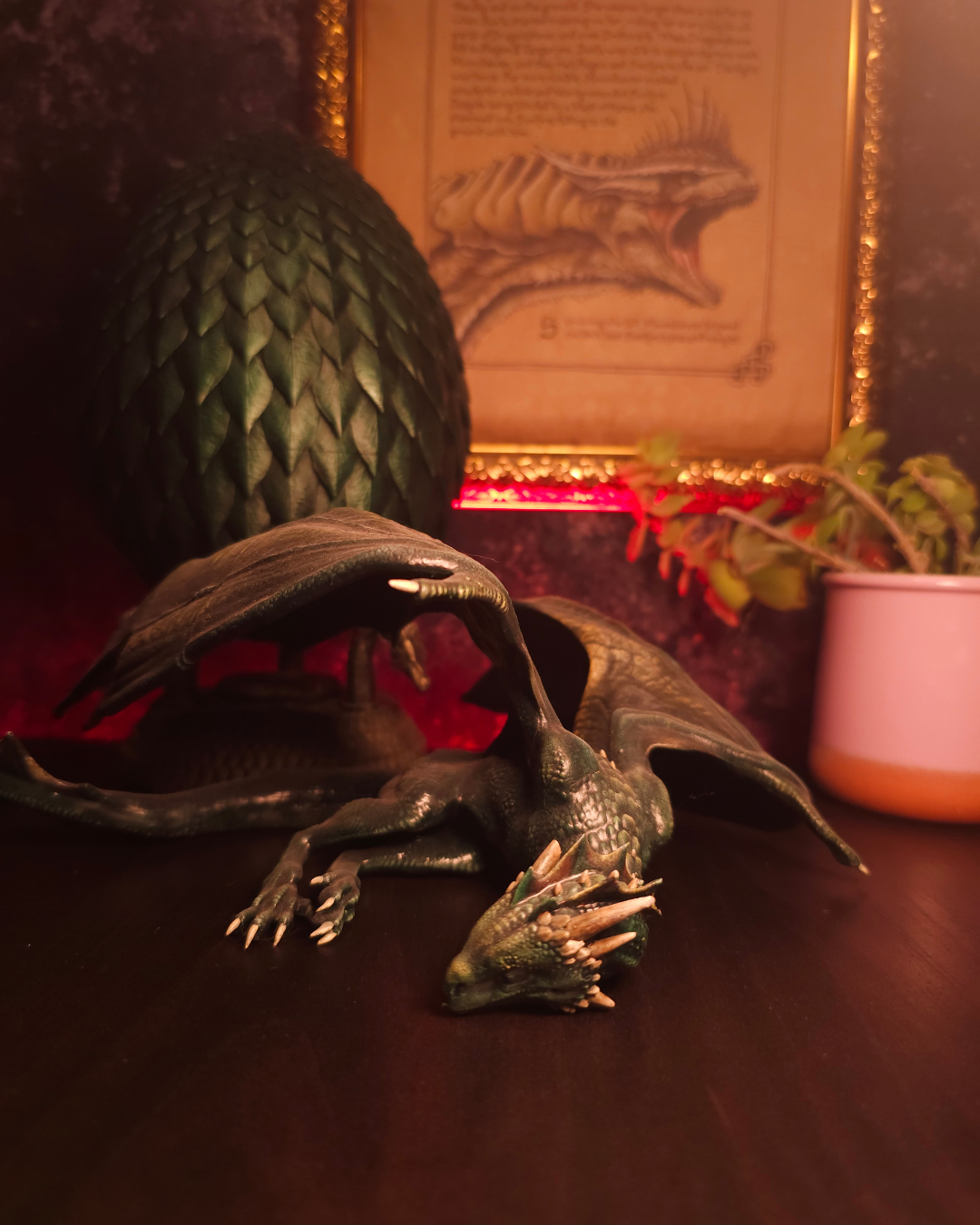 Figurine de dragon vert allongé sur une table, ailes repliées et tête posée au sol, ambiance sombre avec éclairage rouge et décor fantasy en arrière-plan.