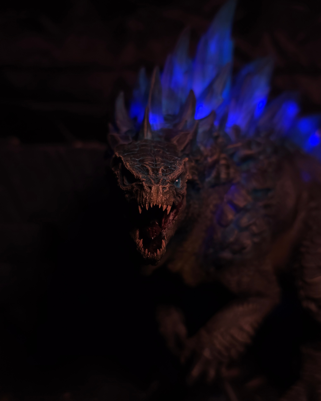 Figurine Godzilla vue de face dans l’ombre, avec crêtes dorsales bleues lumineuses et mâchoire ouverte, créant une scène intense de fantasy dragons et titan collectible.