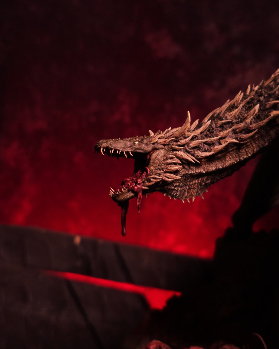Profil du dragon Sheepstealer avec un morceau de chair ensanglanté pendant de sa gueule, éclairé par des teintes rouges pour une ambiance terrifiante.