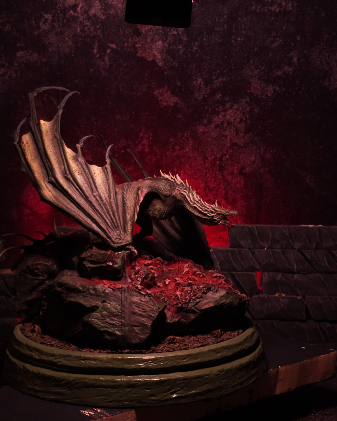 Profil gauche du dragon Sheepstealer dévoilant ses ailes immenses et son corps écailleux, sur un socle rocheux baigné de lumière rouge sang pour un rendu apocalyptique.