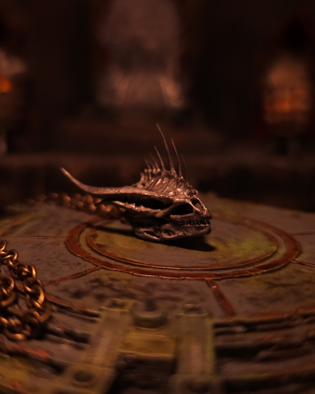 Vue latérale du pendentif représentant le crâne argenté de Moondancer ; cornes fines et arrêtes dorsales acérées, posé sur la table-cartes de Peyredragon sous une lueur mordorée.
