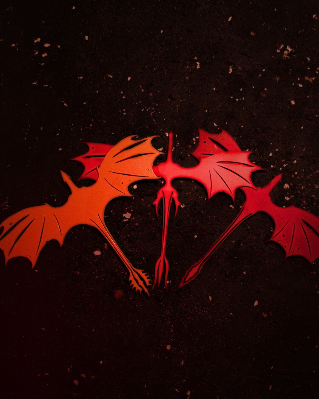 Flat lay of a three-piece bundle of dragon bookmarks in orange, red, and crimson on a dark speckled surface, each with outstretched wings and pointed tails — versatile dragons bookmarks set.