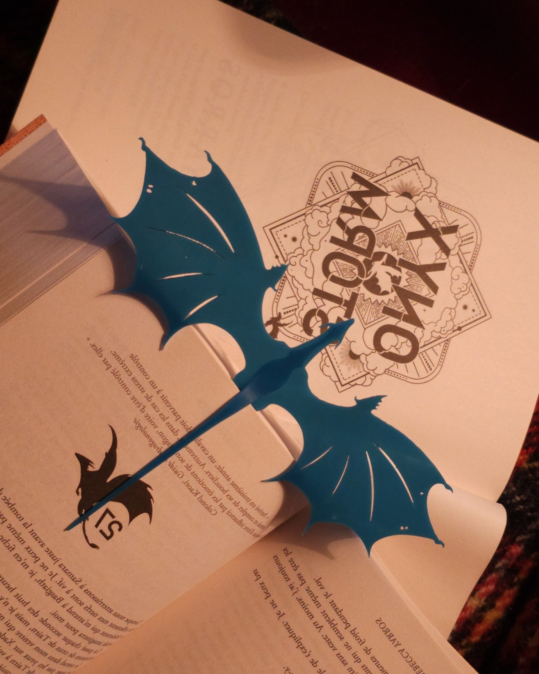 Large marque-page dragon bleu, ailes déployées, inséré dans un livre de fantasy. Accessoire de lecture décoratif rappelant les dragons de Fourth Wing et conçu pour les passionnés de figurines de dragon.