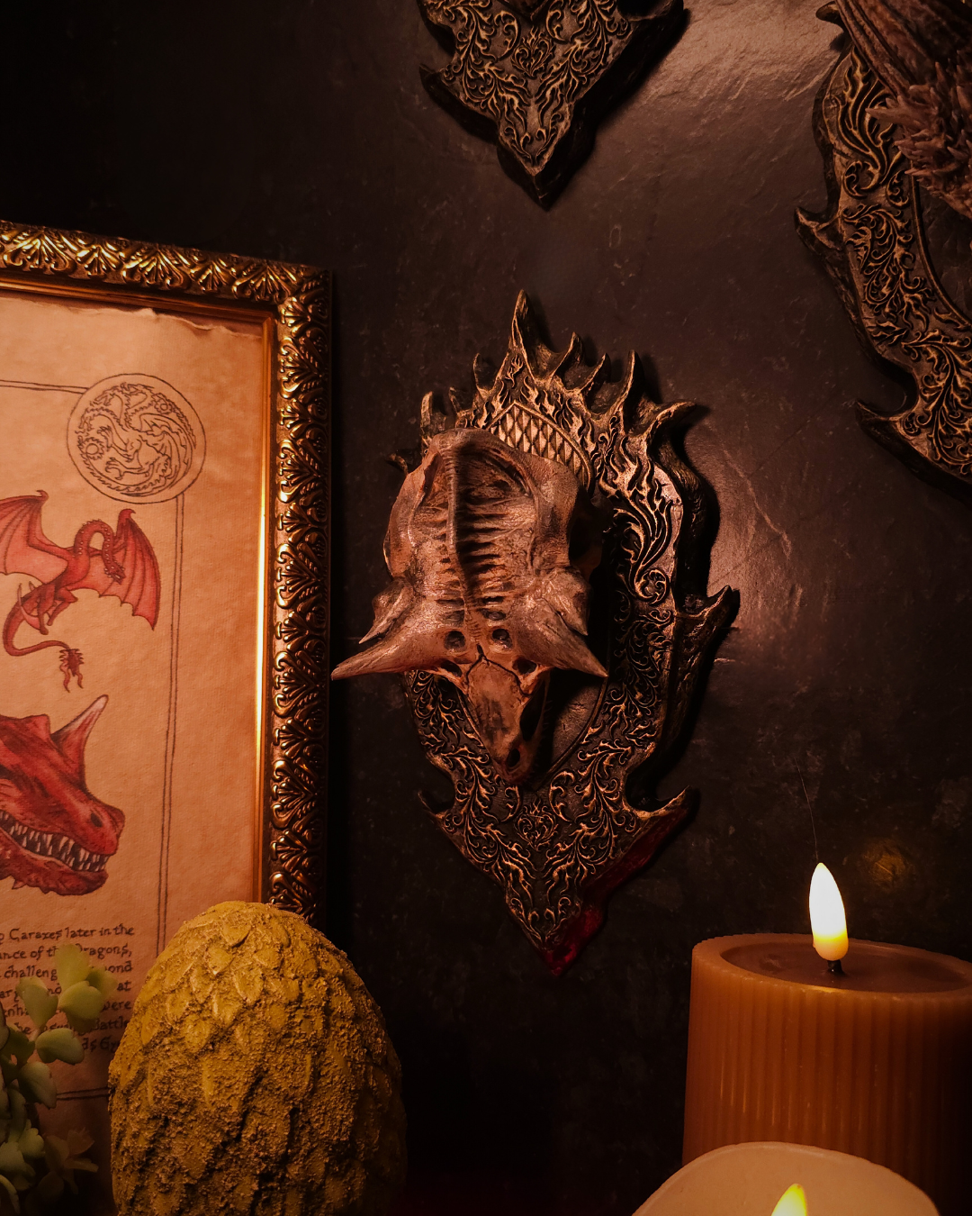 Crâne de dragon Caraxes monté sur plaque murale, placé à côté d’un cadre illustré de dragon et de bougies, dans une scène immersive de fantasy décor gothique.